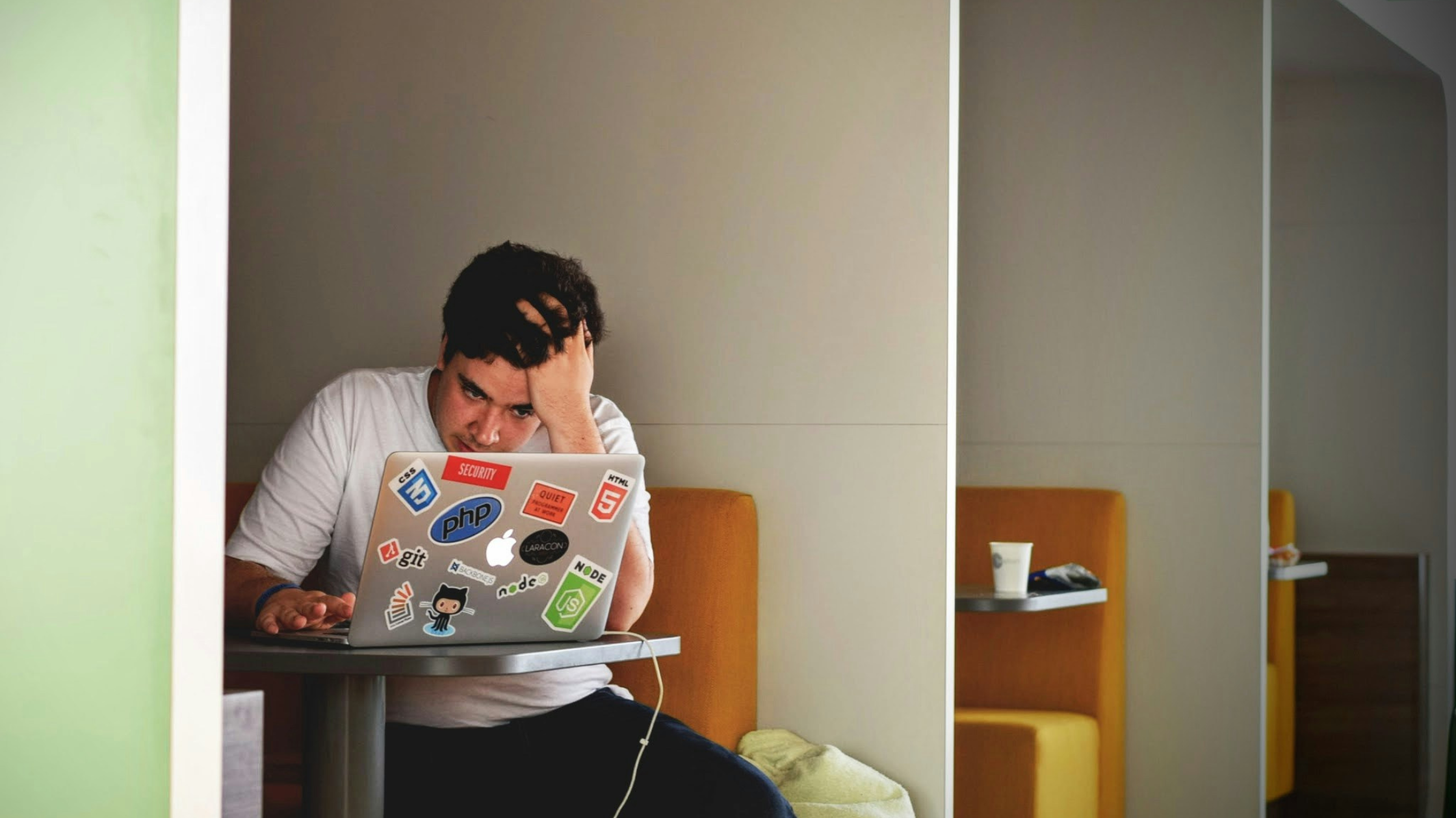 A man works at his laptop in a shared working area. He looks stressed.