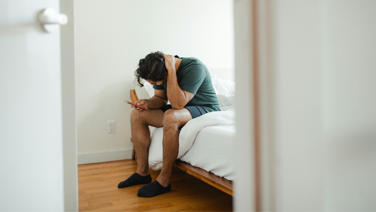 a man sits on the edge of his bed with his phone and looks stressed
