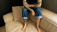 a tattooed woman sits on the edge of her sofa with her arms crossed