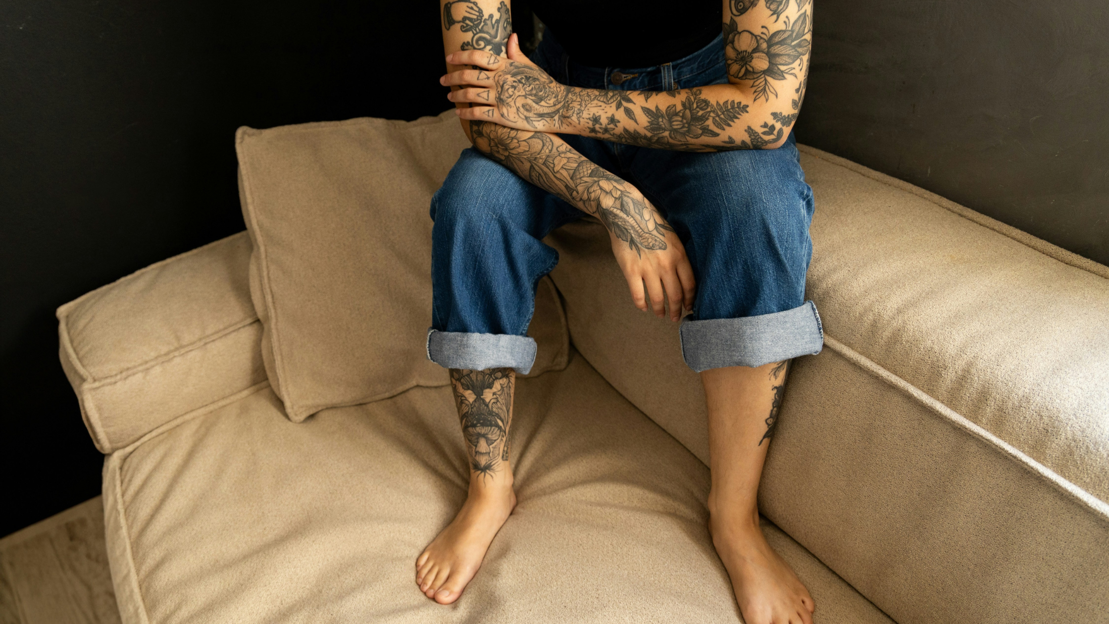 a tattooed woman sits on the edge of her sofa with her arms crossed