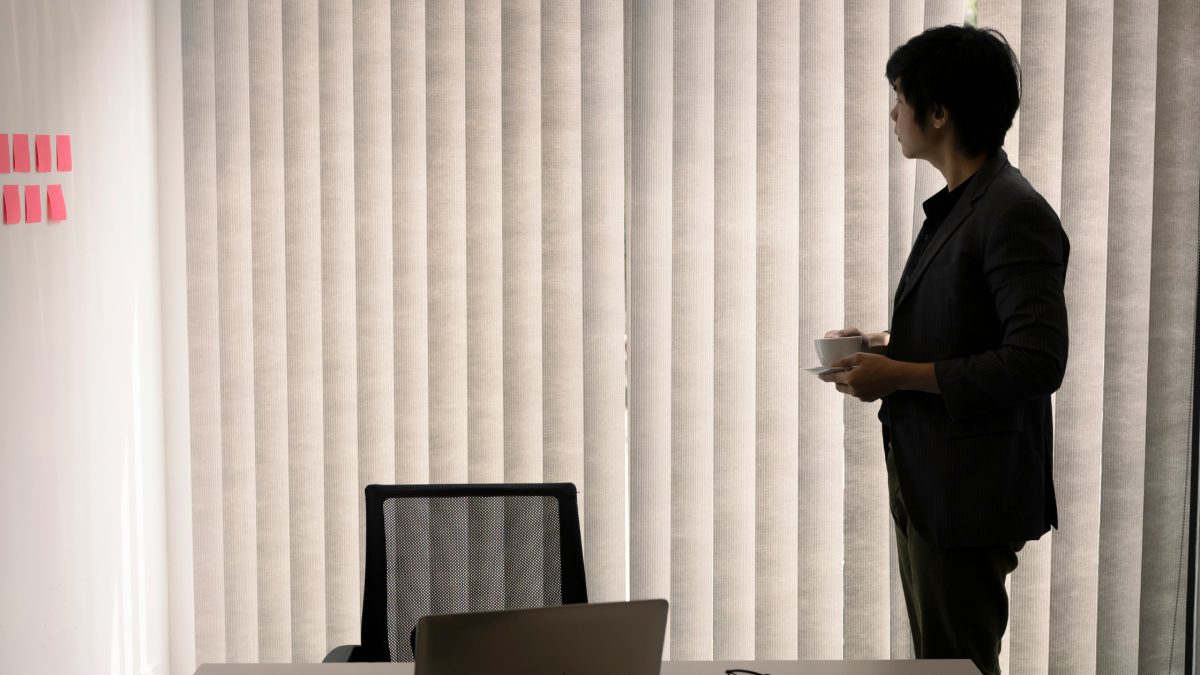 An office worker looks out of his window through the blinds.