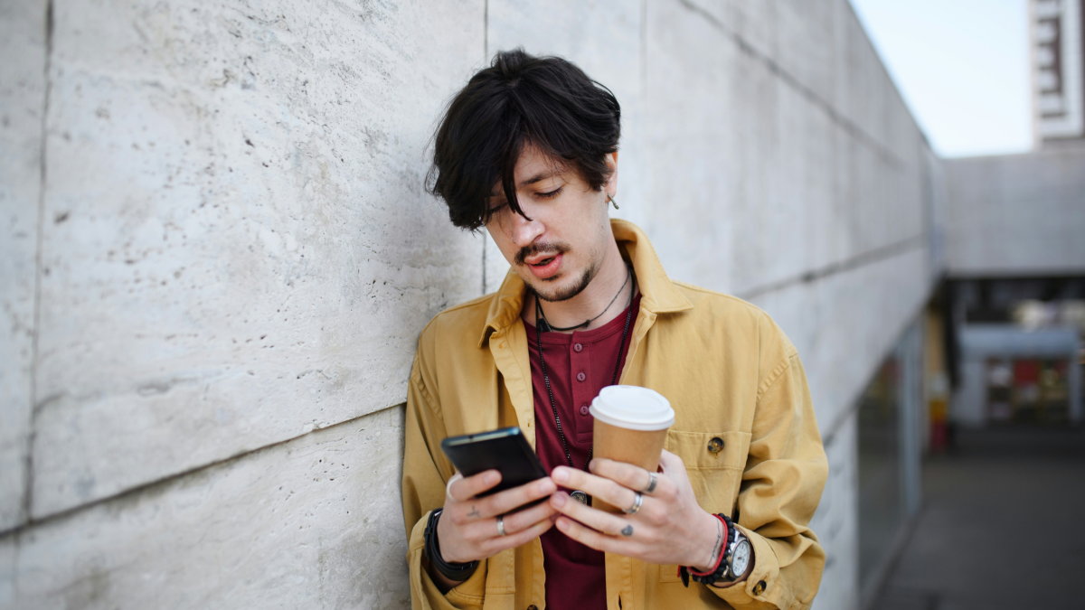 a man leans against a wall with a coffee and uses his phone