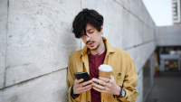 a man leans against a wall with a coffee and uses his phone