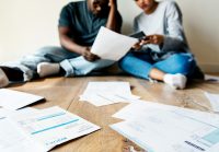A young couple sit on the floor, surrounded by documents, looking worried