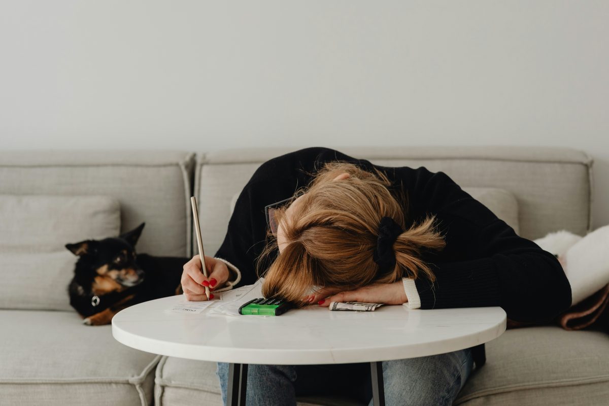 A lady sits with her head on a table, pen in hand