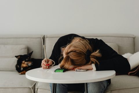 A lady sits with her head on a table, pen in hand